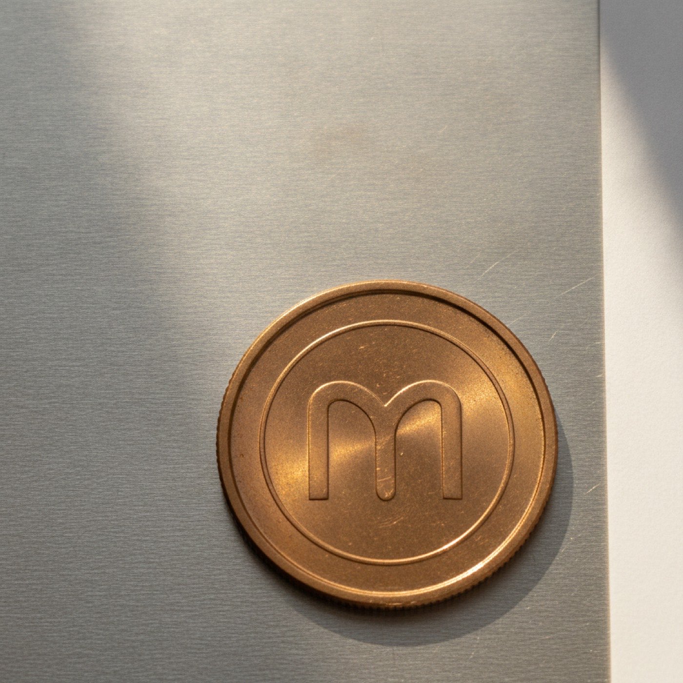 A single Monero coin resting on a brushed aluminium sheet under soft light